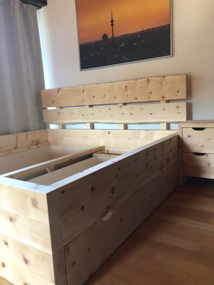 Bettgestell aus hellem Holz mit Kopfteil und Nachtkästchen, an der Wand ein Bild der Münchner Skyline